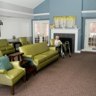 A senior person using a walker sits in a furnished living room with couches, chairs, and a fireplace.