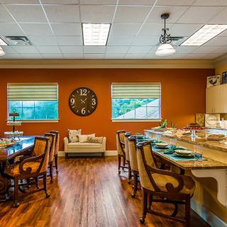 A cozy kitchen and dining area with a warm orange wall, wooden flooring, a central dining table, bar stools at the countertop, and a small sitting area by the window.