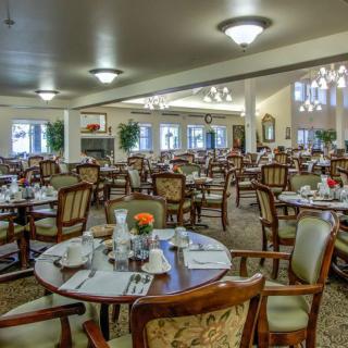 A spacious and elegantly decorated dining room with multiple set tables and chairs, featuring chandeliers and natural lighting.