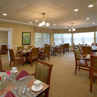 A cozy dining room with neatly arranged tables and chairs, featuring place settings and ample natural light from large windows.