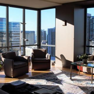 A modern apartment living room with large windows offering a city view, filled with sunlight and stylish furniture.