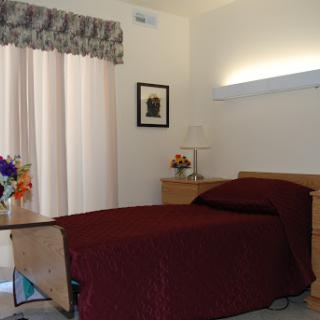 A cozy bedroom featuring a maroon bedspread, curtains, and a small flower arrangement.