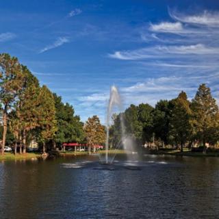 A serene park scene featuring a fountain in the middle of a lake, surrounded by trees and under a blue sky with clouds.