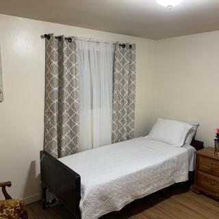 A minimalist bedroom featuring a single bed with white bedding, patterned curtains, and a wooden dresser.