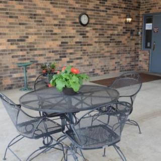 A cozy patio setting with an iron table and chairs against a brick wall, featuring a potted plant and a wall-mounted clock.