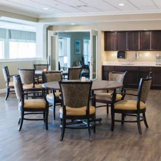 A well-lit dining area with tables and chairs, featuring wood flooring and a kitchenette in the background.