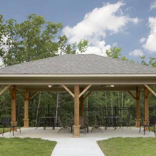A wooden gazebo with a shingled roof, surrounded by trees under a partly cloudy sky.