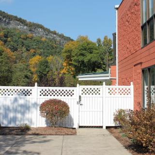 A view of a white fence and gate beside a brick building, with colorful autumn trees in the background.