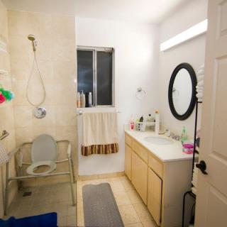 A well-lit bathroom featuring a shower area with safety chair, a vanity with a round mirror and sink, and neatly arranged toiletries.