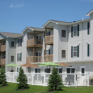 A modern apartment building with multiple balconies and a fenced outdoor area, surrounded by grass and trees.