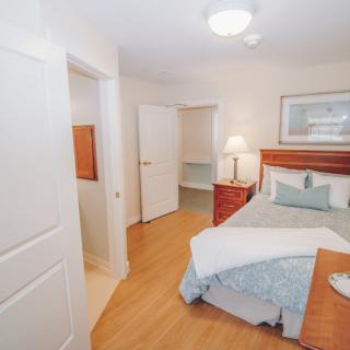 A cozy bedroom with wooden flooring, featuring a bed, a bedside lamp, and artwork on the wall.
