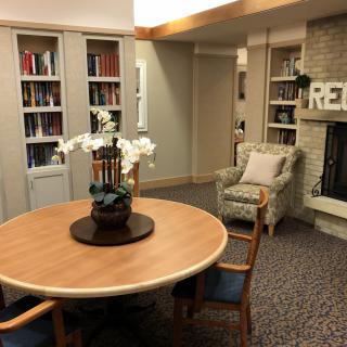 A cozy library reading room with a wooden table, surrounded by shelves filled with books and a fireplace.
