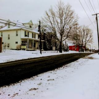 A quiet winter scene featuring snow-covered ground, bare trees, and suburban homes lining the road.