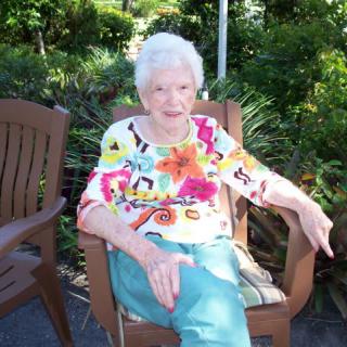 An elderly woman sitting outdoors in a garden, wearing a colorful shirt and enjoying a sunny day.