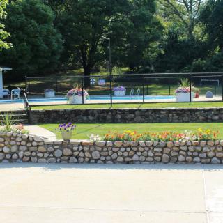 A fenced swimming pool area surrounded by greenery and a stone wall.