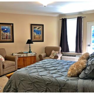 A cozy bedroom featuring a large bed with a blue comforter, two armchairs, decorative wall art, and a window with curtains.