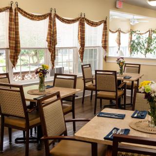 A cozy dining area with neatly arranged tables and chairs, adorned with flowers and bathed in natural light from the windows.