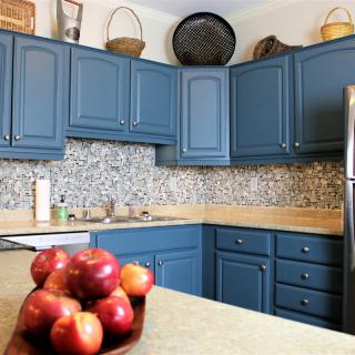 A cozy kitchen with blue cabinets, a fruit bowl filled with apples on the countertop, and a tiled backsplash.