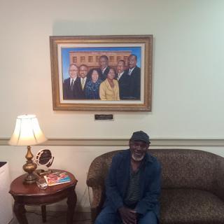 A man sitting on a couch in a room with a portrait of a group of people hanging on the wall.