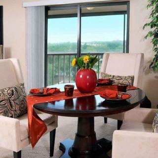 A cozy dining space with a round table elegantly set with a red vase and matching tableware, overlooking a scenic view through glass doors.