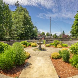 A serene garden space featuring a central fountain, surrounded by lush greenery and a clear blue sky.