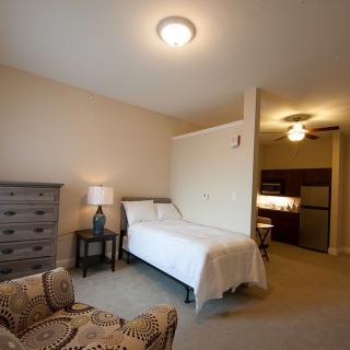 A cozy bedroom with a single bed, dresser, and chair, featuring neutral tones and warm lighting.