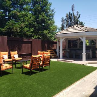 A cozy backyard with a gazebo, outdoor seating on artificial grass, surrounded by trees and a fence.