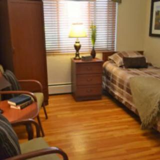 A cozy bedroom with wooden flooring, featuring a bed, a bedside table with a lamp, and chairs.
