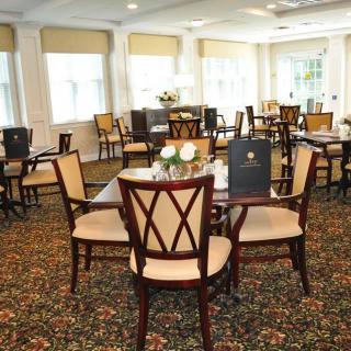 A well-decorated dining area with multiple tables and chairs, featuring elegant carpeting and large windows providing ample natural light.