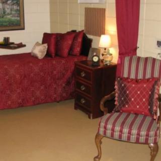 A cozy bedroom with a red and beige color scheme featuring a bed, patterned chair, nightstand, and lamp.