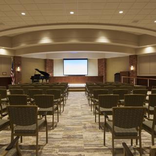 A spacious auditorium with rows of chairs facing a stage equipped with a grand piano and a projector screen.