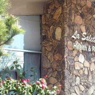 The image shows the exterior stone facade of a healthcare facility with visible signage and surrounding plants.