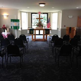 A small chapel interior with seating arranged facing an altar, flanked by windows and flags.
