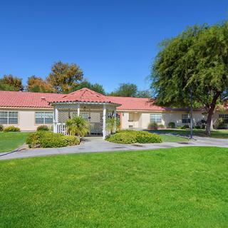 A sunny day in a suburban neighborhood, featuring a residential building with a lush green lawn and trees.