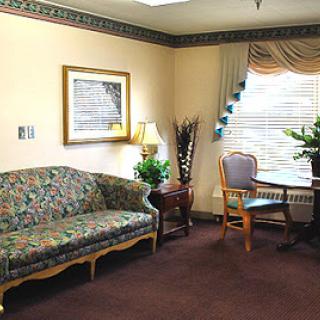 A cozy living room setup featuring a floral patterned sofa, a wooden table and chair, along with decorative plants and wall art.
