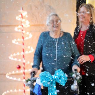 An elderly woman with a walker, decorated for the holidays, is accompanied by a caregiver in a festive setting with lights and snow.