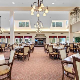 A spacious dining hall with multiple tables and chairs, featuring elegant chandeliers and a carpeted floor.