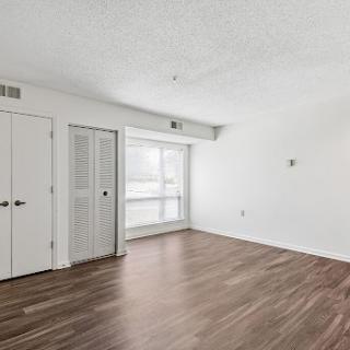 A spacious and empty interior room with white walls and wooden flooring, illuminated by natural light from the windows.