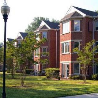 A row of brick apartment buildings with well-maintained lawns and trees in a suburban setting.