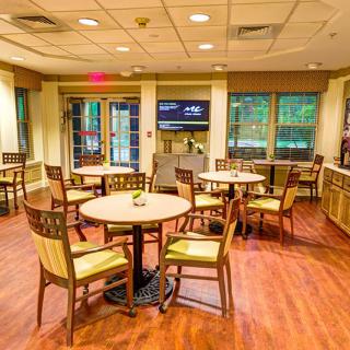 A cozy dining area with tables and chairs arranged on a wooden floor, featuring large windows for natural light.