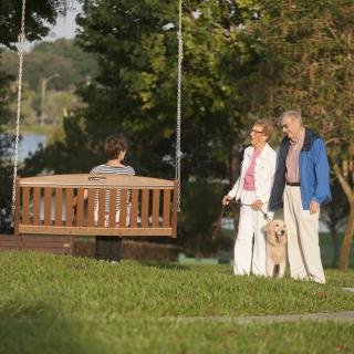 A couple walking their dog in a park, passing by a person sitting on a swing.