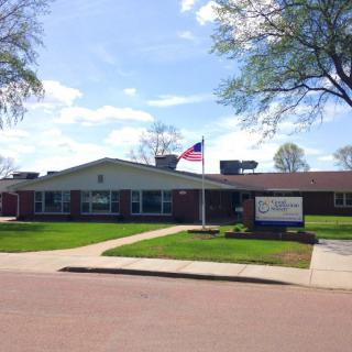A single-story brick building with an American flag flying, surrounded by a green lawn and trees on a sunny day.