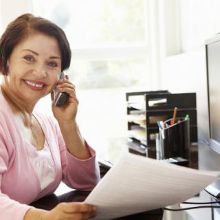 A smiling woman engaged in a phone conversation while reviewing paperwork in her office.