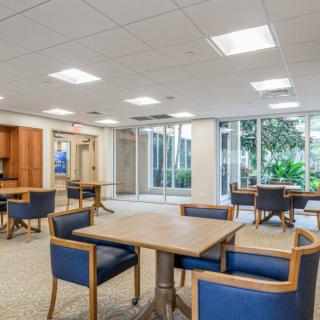 A modern office lounge with tables and chairs, featuring large windows and natural lighting.