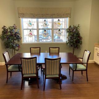 A cozy dining area featuring a wooden table surrounded by six chairs, with large windows and decorative plants on either side.