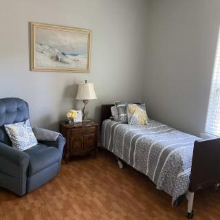A cozy bedroom with a single bed, blue armchair, nightstand, lamp, and a painting on the wall.