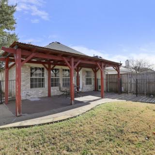 A backyard with a covered patio featuring a pergola, surrounded by a lawn and fenced area.