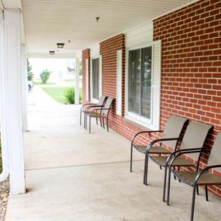 A covered porch with several chairs lined up against a brick wall.