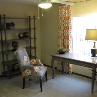 A cozy home office with a desk, upholstered chair, and shelving unit near a window with patterned curtains.