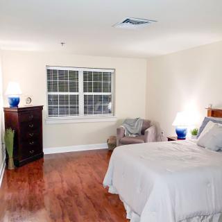 A cozy bedroom with wooden floors, featuring a bed, dresser, and armchair near a bright window.